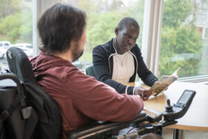 An advisor wearing a black top goes over notes with young student in a power chair wearing a red sweatshirt